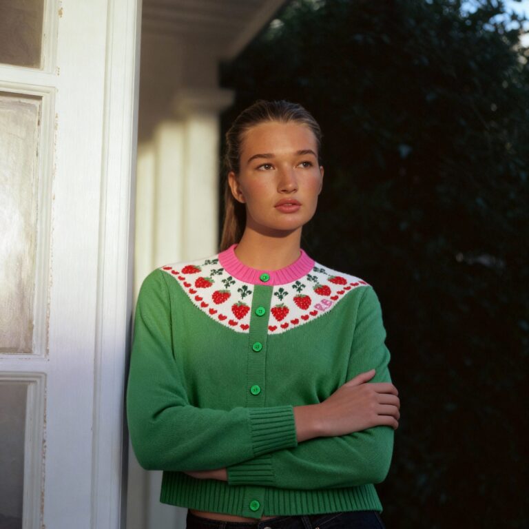 Model in a green strawberry-pattern cardigan, arms crossed, leaning against a doorframe in soft daylight